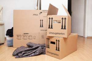 A collection of moving supplies in a room, including cardboard boxes and moving blanket on light hardwood floor with a plastic bin peeking out from behind the largest cardboard box