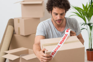 Young man packing up and wrapping a box with tape that says "fragile" in red